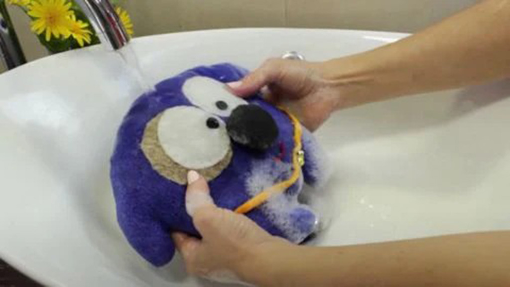 Hands gently washing a purple plush toy with soap under running water in a white sink.