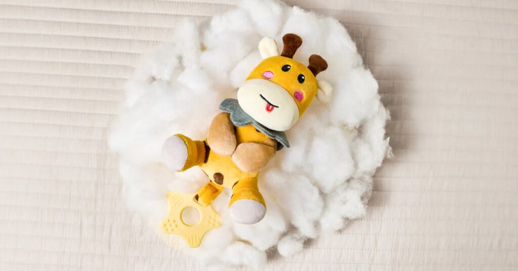 A small yellow giraffe plush toy with a teether attached lies on a soft pile of white stuffing fiber, placed on a light-colored quilted surface.