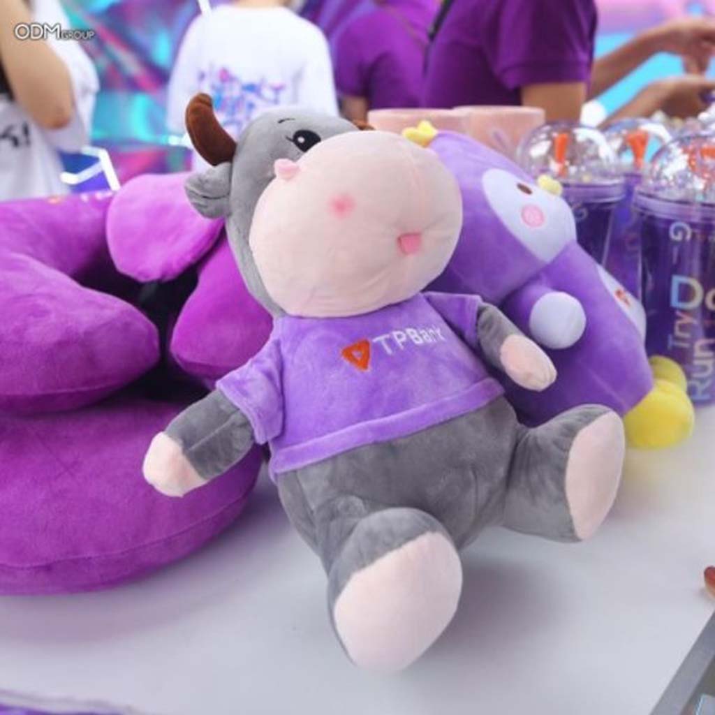 A gray cow plush toy wearing a purple TPBank shirt displayed on a table with other purple promotional items during a marketing event.