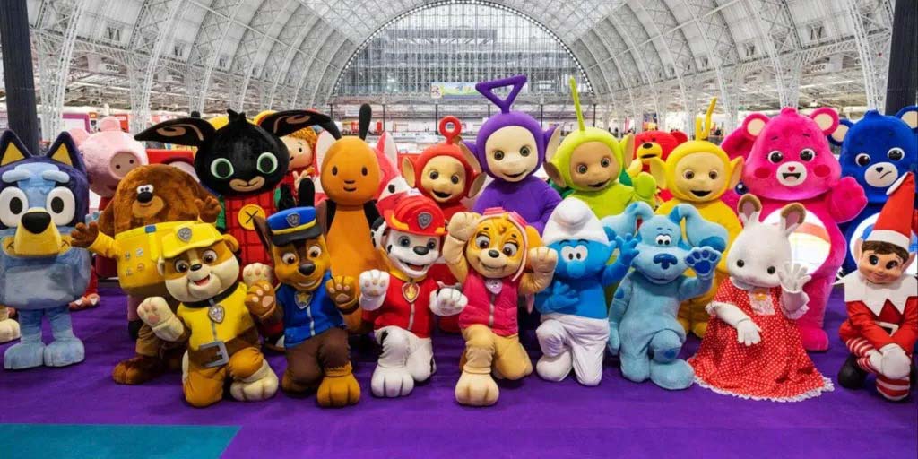 Large group of colorful mascot characters—including Paw Patrol, Teletubbies, Bluey, Care Bears, and others—posing together inside a spacious exhibition hall.