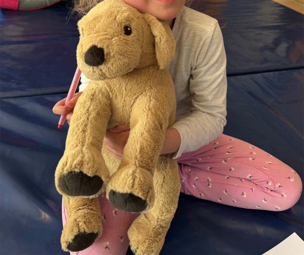 Young child sitting on a blue mat, holding a soft beige stuffed dog with dark paws and nose while smiling gently.