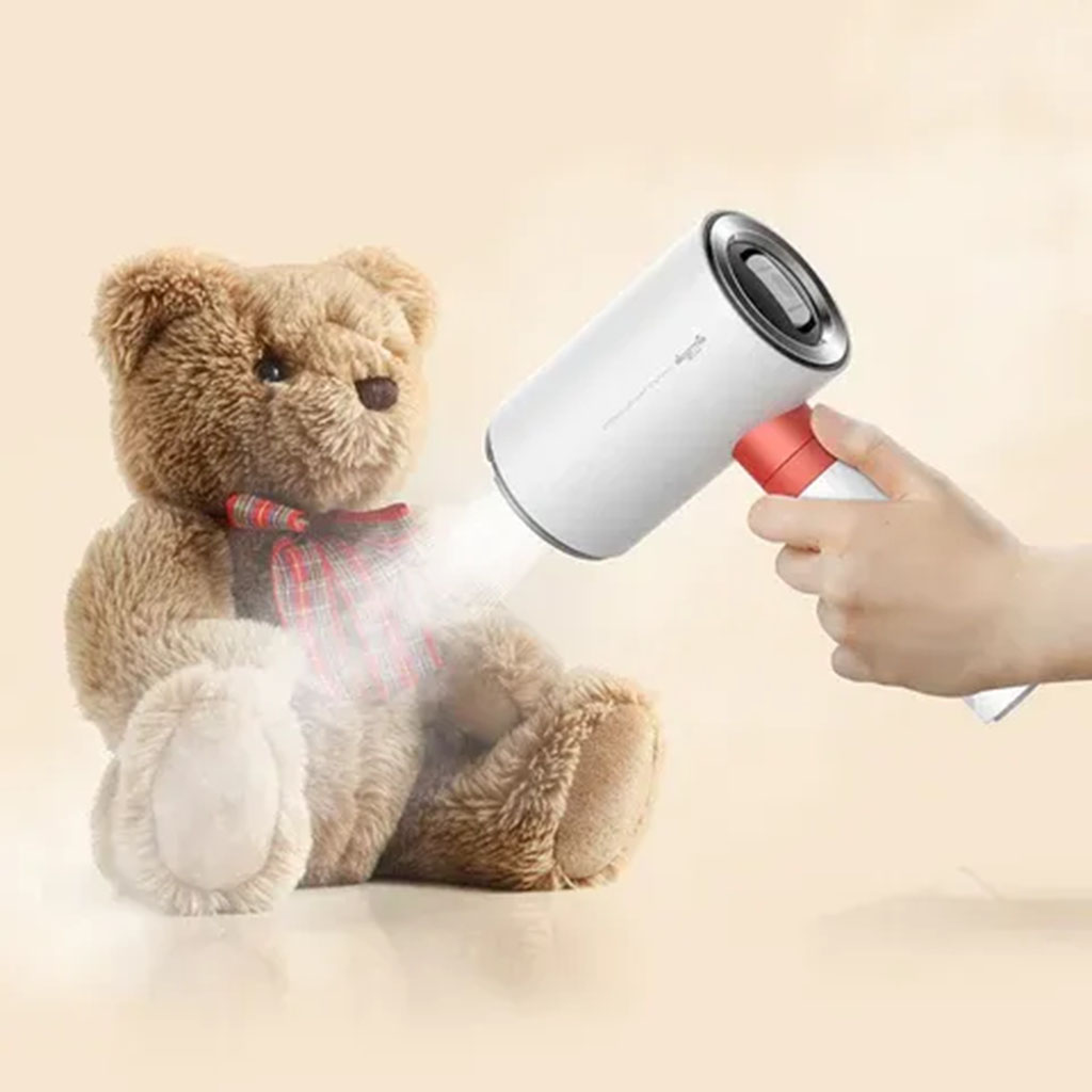Hand using a white garment steamer to sanitize and refresh a brown teddy bear with a red plaid bow, demonstrating plush toy cleaning care.