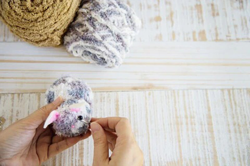Hands attaching facial details to a small handmade amigurumi plush using yarn, with soft yarn balls in the background on a wooden table.