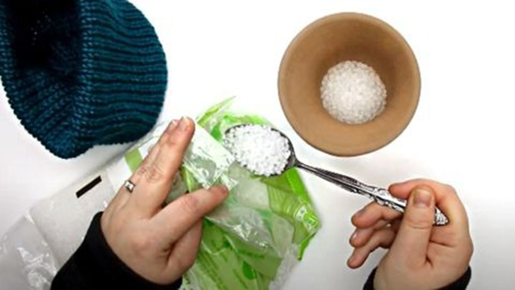 Hands scooping small white poly pellets with a spoon into a plastic bag, with a bowl of pellets and fabric item nearby.