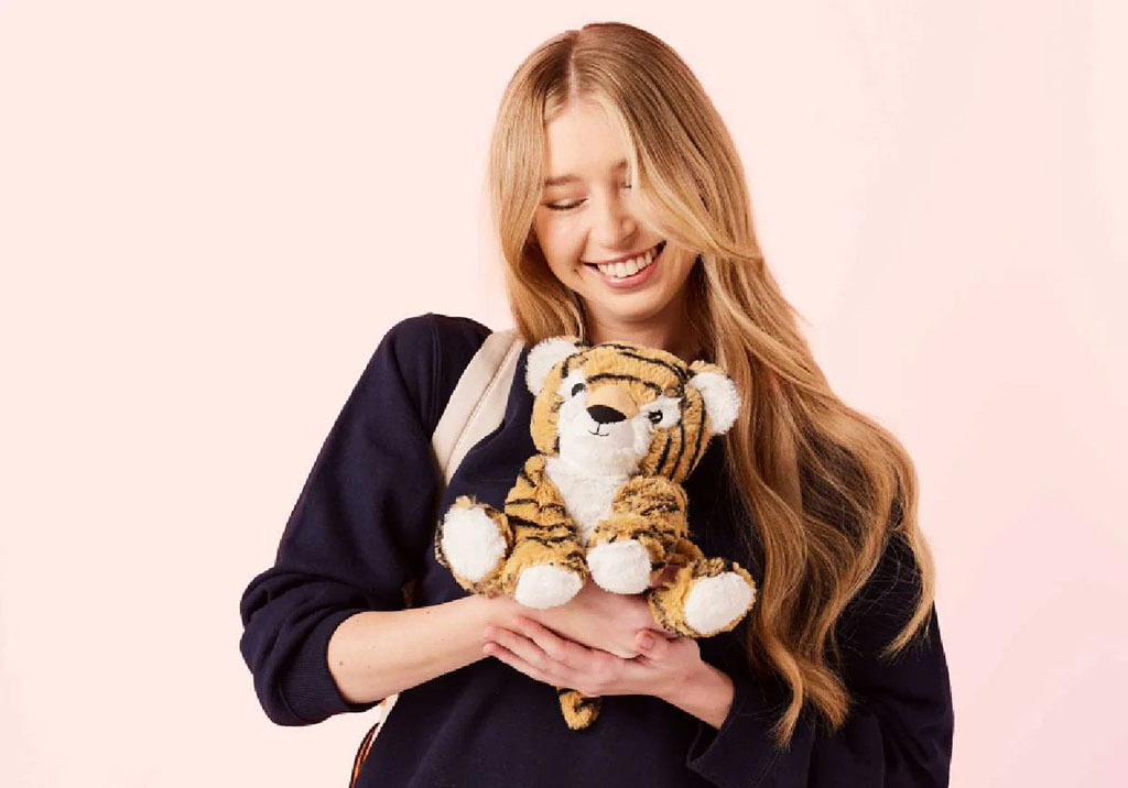 A young woman with long blonde hair smiles while hugging a small plush tiger toy against a soft pink background.