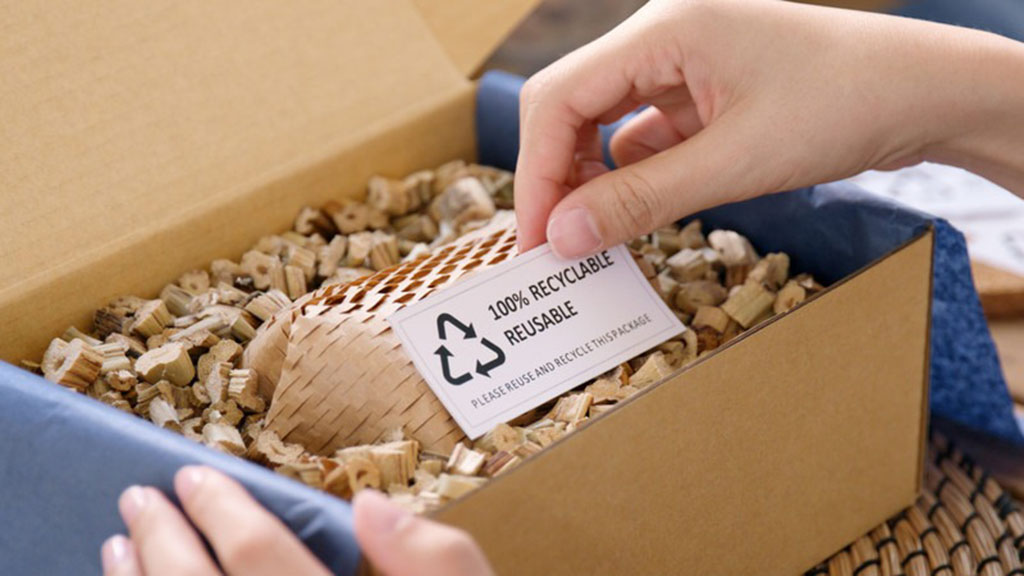 Hands opening a cardboard box filled with biodegradable paper cushioning, featuring a label that says “100% recyclable reusable,” showing eco-friendly packaging materials for sustainable shipping.