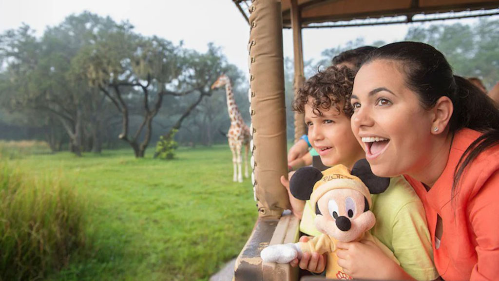 Smiling woman and child holding a Mickey Mouse plush toy while watching a giraffe during a safari ride at Disney’s Animal Kingdom.