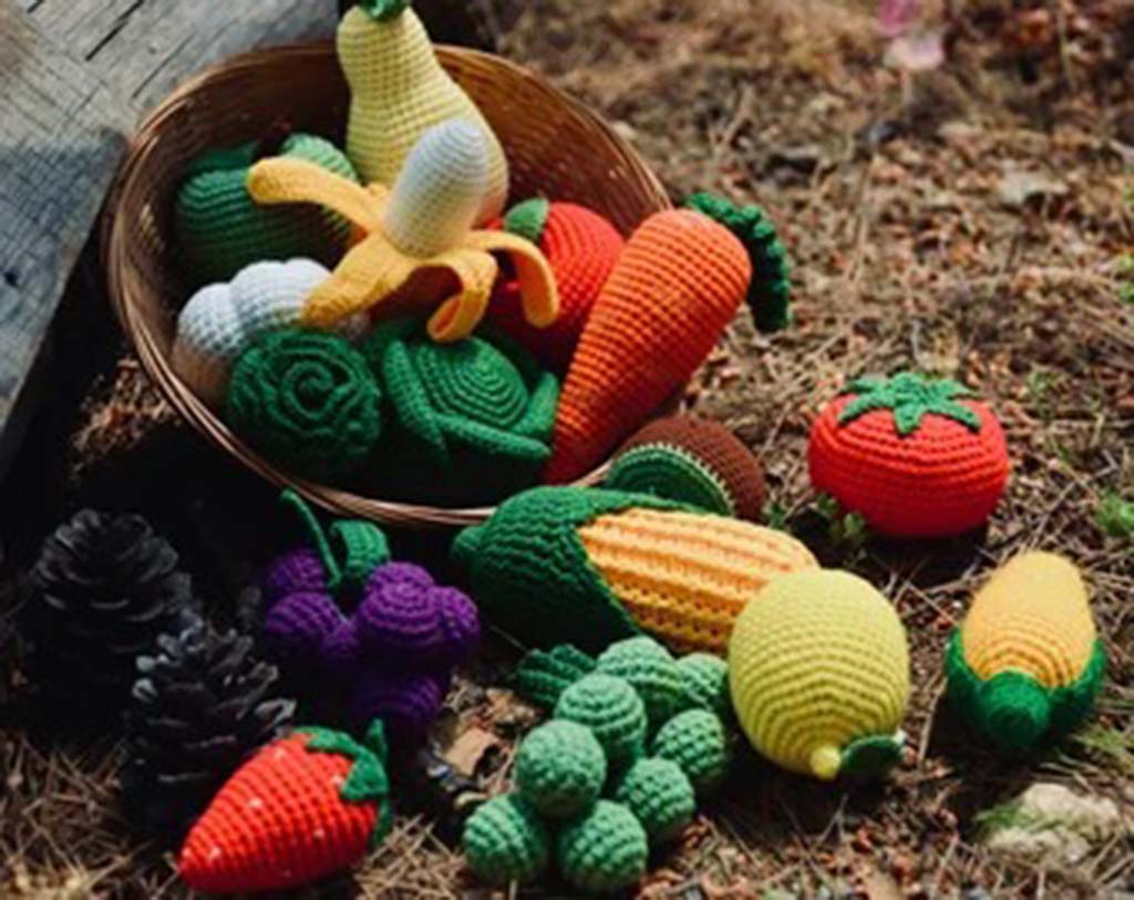 A basket of colorful crochet fruits and vegetables, including a banana, carrots, lettuce, grapes, corn, tomatoes, strawberries, and lemons, arranged outdoors on the ground.