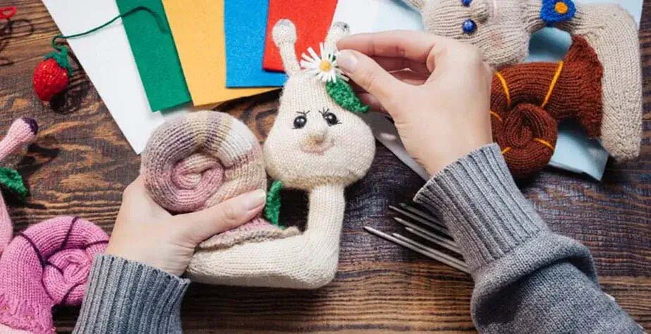 Hands adding a flower decoration to a knitted snail plush toy surrounded by felt sheets, yarn, and crafting tools.