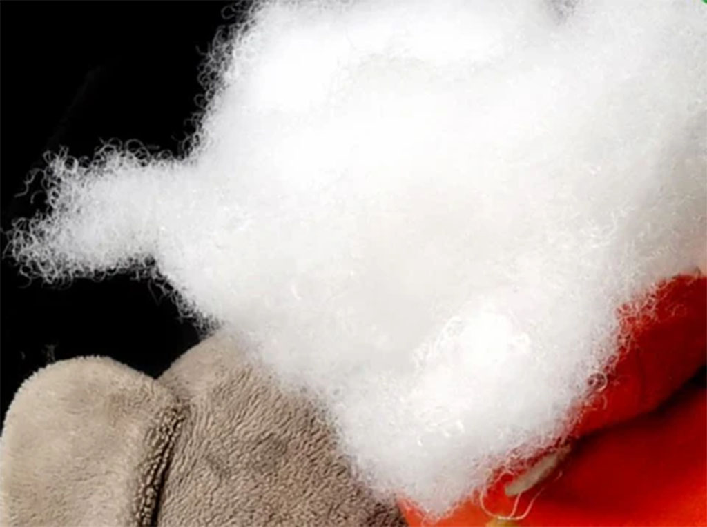 Close-up of soft white polyester fiberfill used as stuffing for plush toys, shown resting on fabric with visible fluffy, curly fibers.