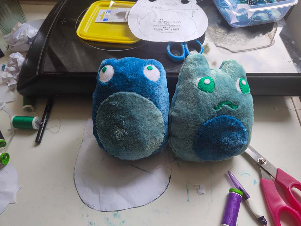 Two small handmade blue plush creatures on a crafting table surrounded by thread, scissors, and pattern pieces.