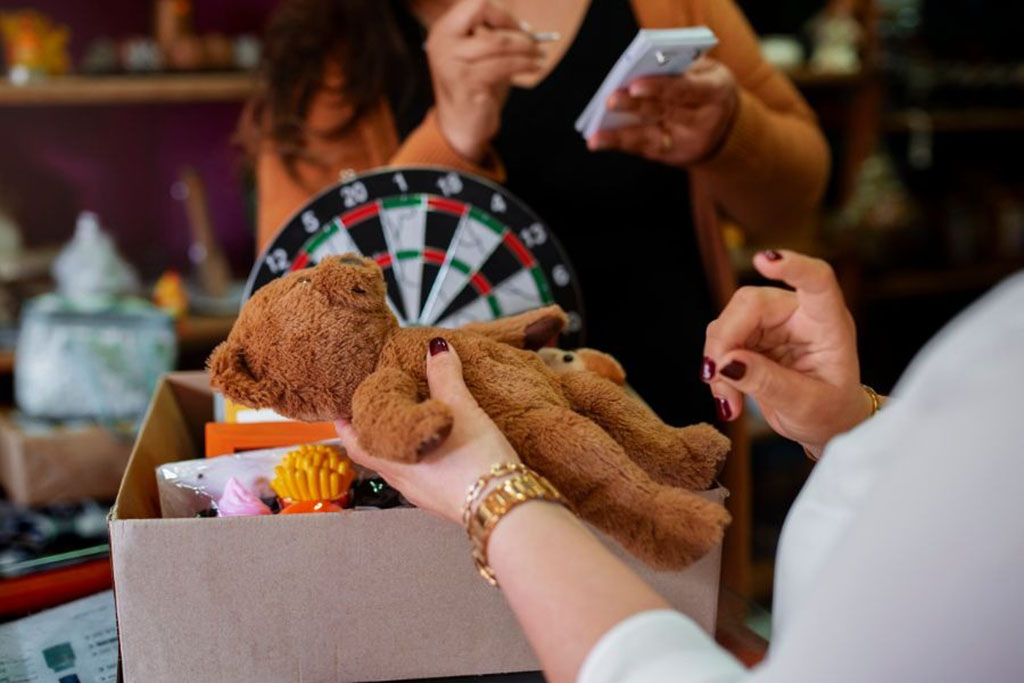 A person holding a brown teddy bear for inspection while another person takes notes, with a dartboard and various items visible in the background.