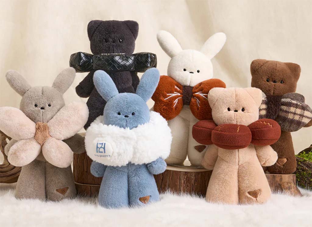 A group of soft plush bunny toys in various colors, each wearing a decorative scarf or collar, displayed on a fluffy surface with wooden props in the background.
