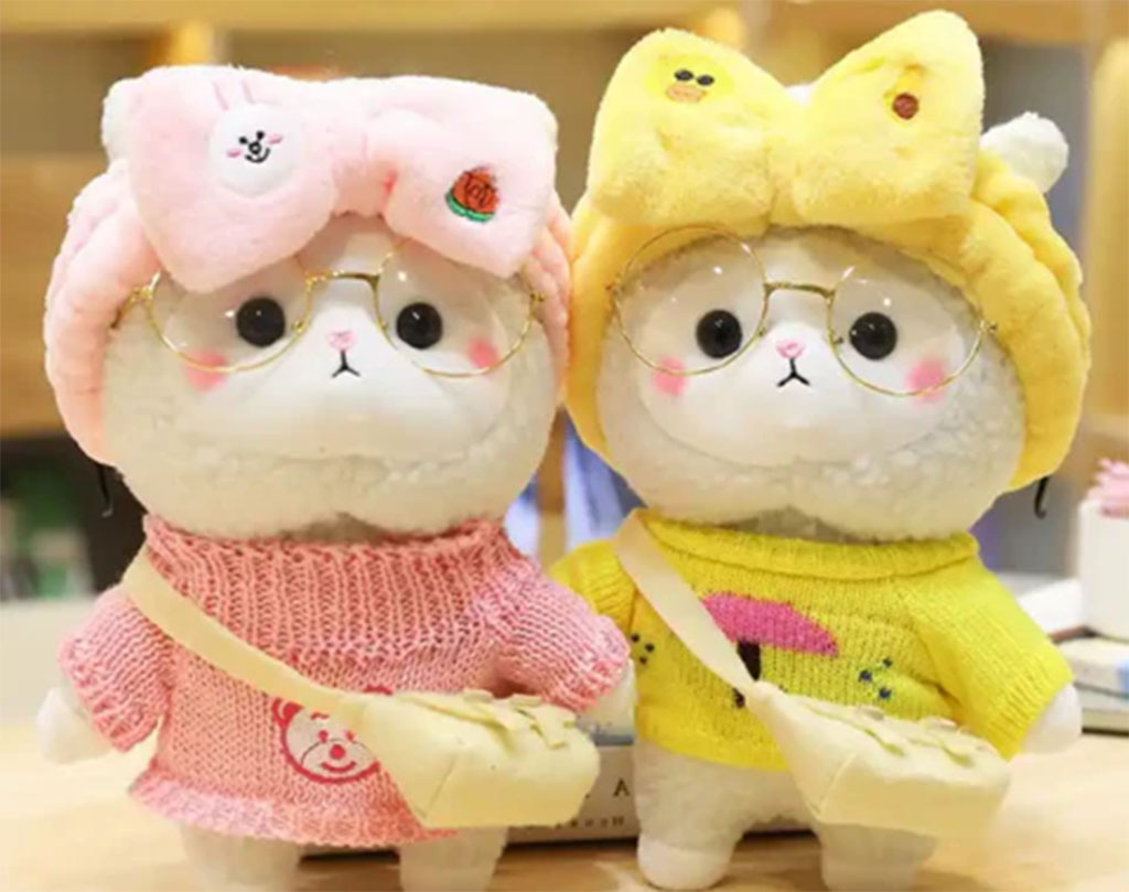 Two adorable plush cat dolls wearing glasses, colorful sweaters, and matching bows — one in pink and one in yellow — each carrying a small fabric bag.