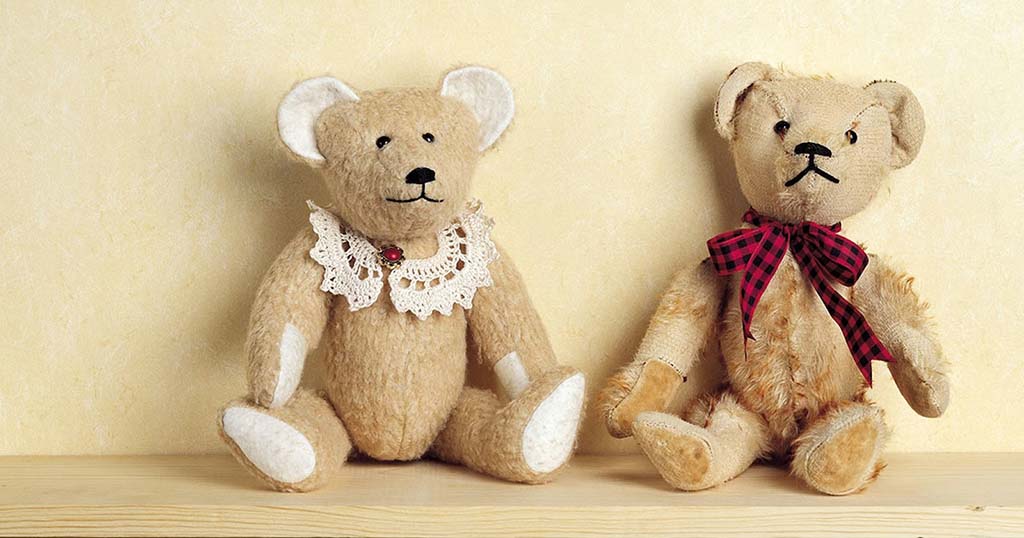 Two classic teddy bears sitting on a wooden surface, one wearing a lace collar and the other with a red plaid ribbon, representing early handmade teddy bear styles.