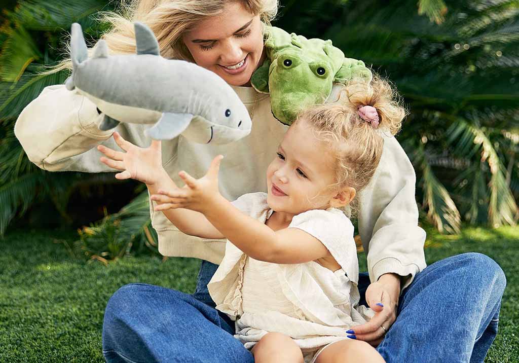 Mother and young daughter playing outdoors with shark and frog plush toys, smiling and reaching toward each other.