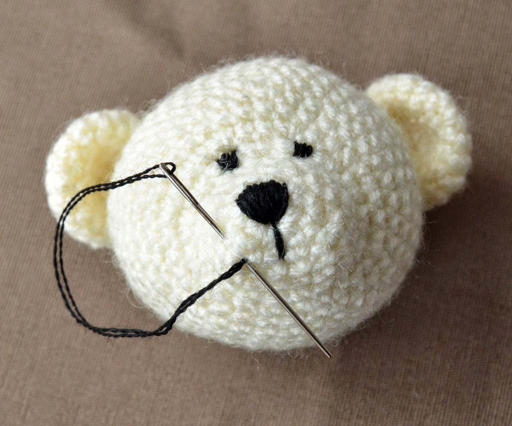 A crocheted bear head with a needle and black thread being used to stitch its facial details.