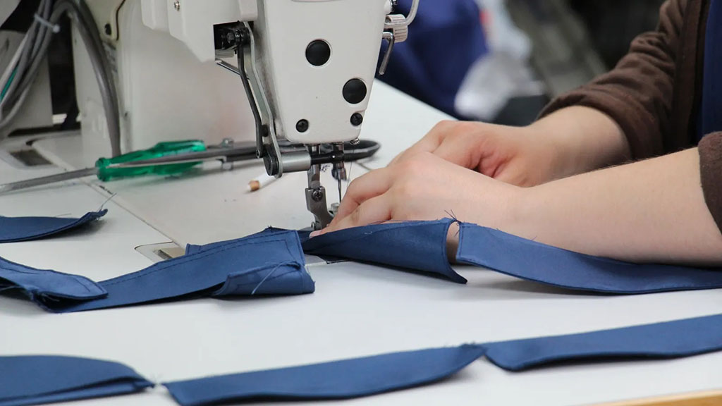 Close-up of a worker sewing blue fabric strips using an industrial sewing machine, with hands guiding the material on a white workstation.