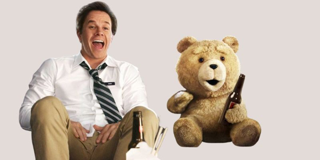  A man sitting and laughing next to a talking teddy bear holding a bottle, set against a neutral background.