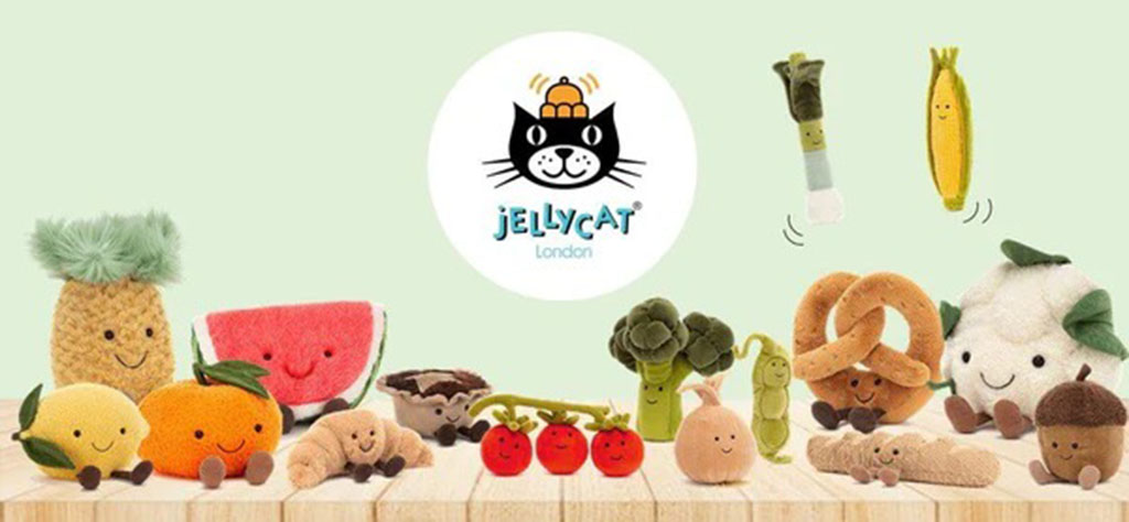 A collection of Jellycat food-themed plush toys, including smiling fruits, vegetables, and snacks such as pineapple, watermelon, broccoli, peas, pretzel, and cauliflower, arranged on a wooden surface with the Jellycat London logo centered above them.