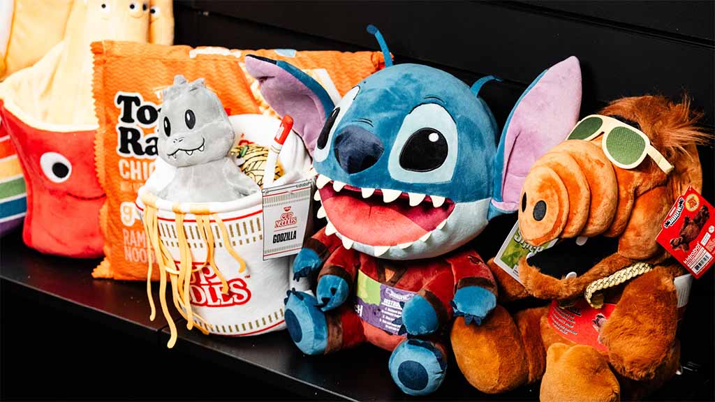 Colorful assortment of character plush toys, including Stitch, Godzilla in a noodle cup, ALF, and other themed plush items on a display shelf.