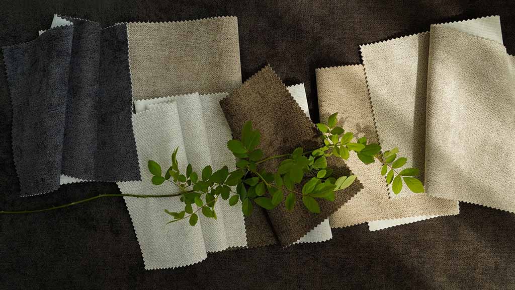 A collection of neutral-toned fabric swatches in grays, beiges, and browns arranged on a dark surface with a green leafy branch placed on top.
