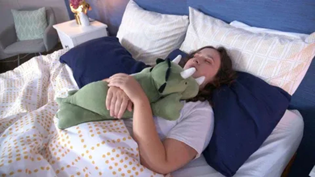 Person sleeping in bed while hugging a green weighted dinosaur plush toy for relaxation and comfort.