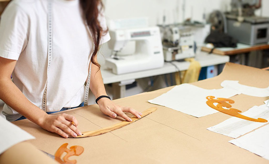 Designer drawing plush toy patterns on brown paper using rulers in a sewing workshop with fabric pieces and sewing machines in the background.