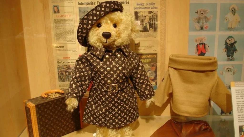 A teddy bear dressed in a luxury monogram coat and hat, displayed beside a matching suitcase and clothing items in a museum-style showcase.