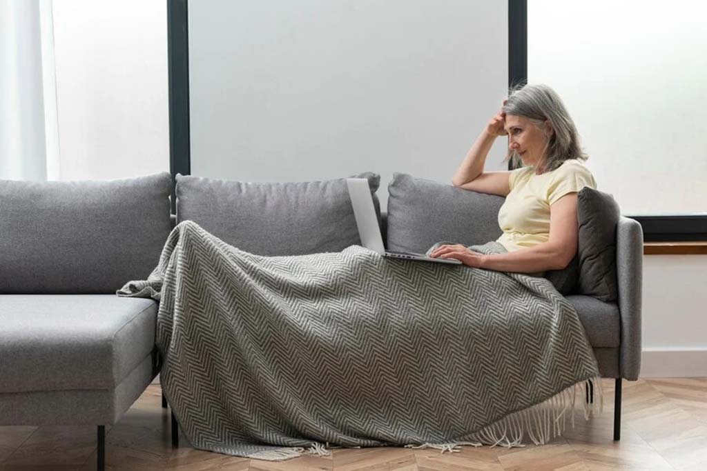 A woman sitting on a gray sofa, covered with a patterned throw blanket, working on her laptop in a bright living room.