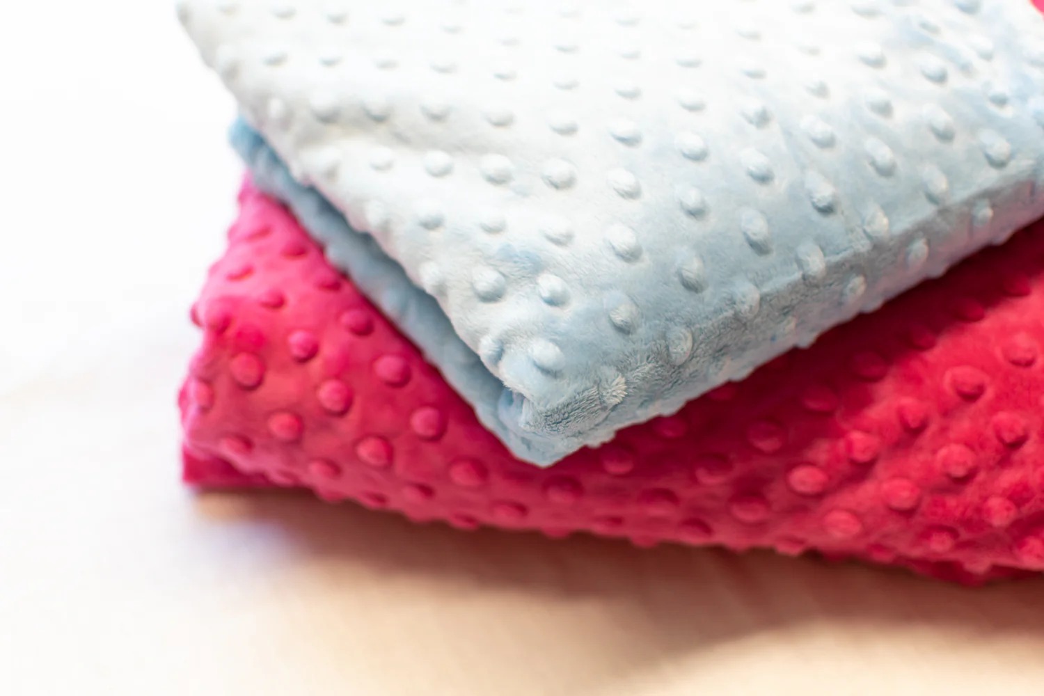 Close-up of folded blue and pink minky dot fabrics with raised textures, showing the plush softness commonly used in baby blankets and plush toys.