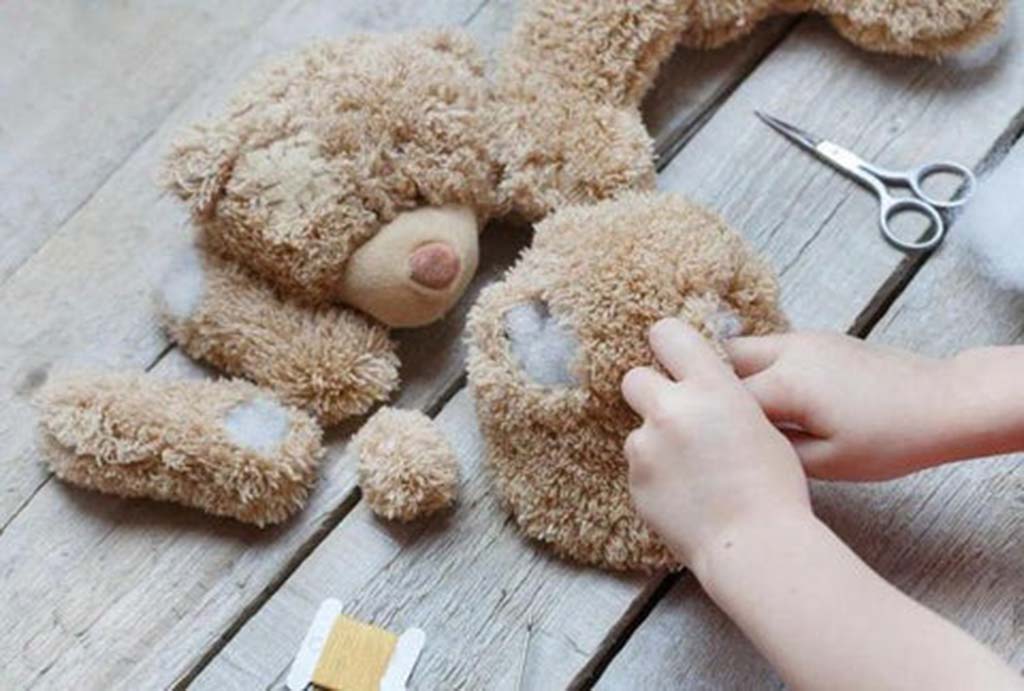 Close-up of hands sewing and restuffing a brown teddy bear on a wooden table, with scattered stuffing, thread, and scissors nearby.