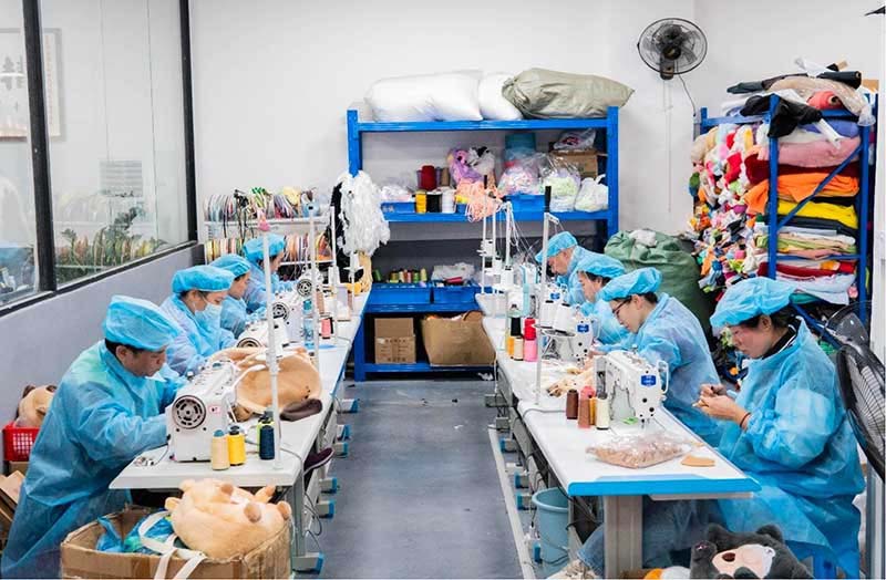 Plush toy factory sewing department with workers in blue protective uniforms operating industrial sewing machines, surrounded by fabric shelves, stuffing materials, and plush components during stuffed animal production.