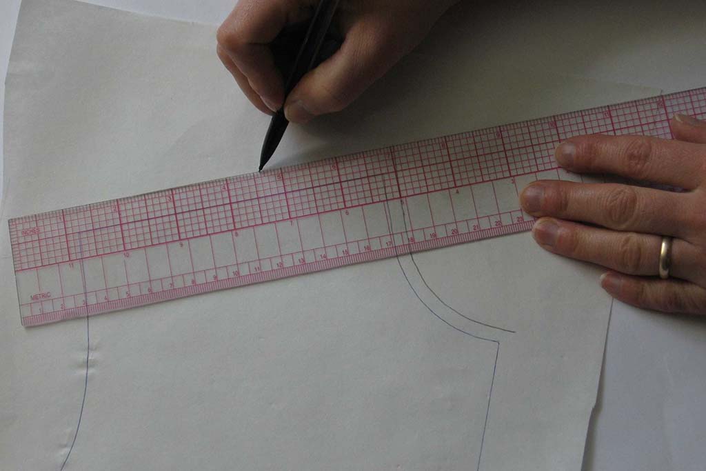 Hands using a transparent gridded ruler to draw precise seamlines on pattern paper for sewing or plush toy pattern drafting.