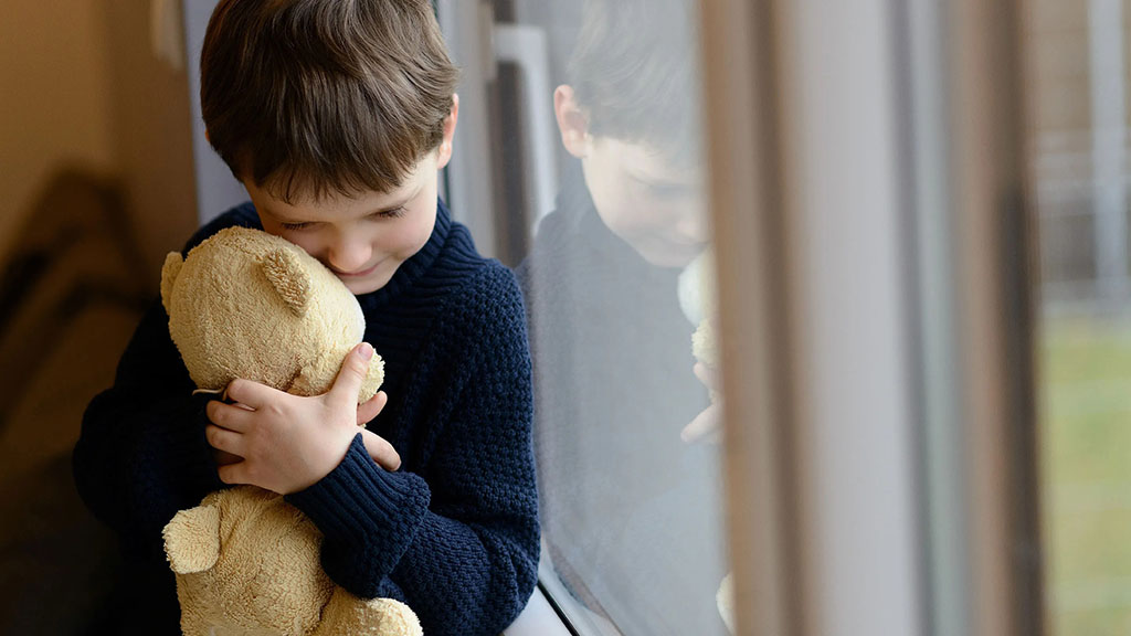 A young child warmly hugging a soft teddy bear while standing near a window, showing the comforting and emotional connection kids have with plush toys.