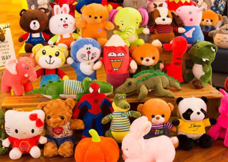 A large assortment of colorful plush toys, including animals, cartoon characters, and fantasy creatures, arranged on wooden shelves in a bright indoor setting.