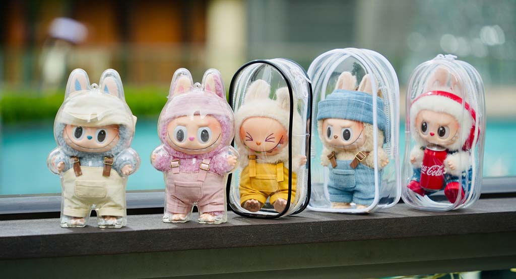 Five cute bunny-themed plush dolls dressed in colorful outfits, each displayed in a transparent travel case by a poolside.