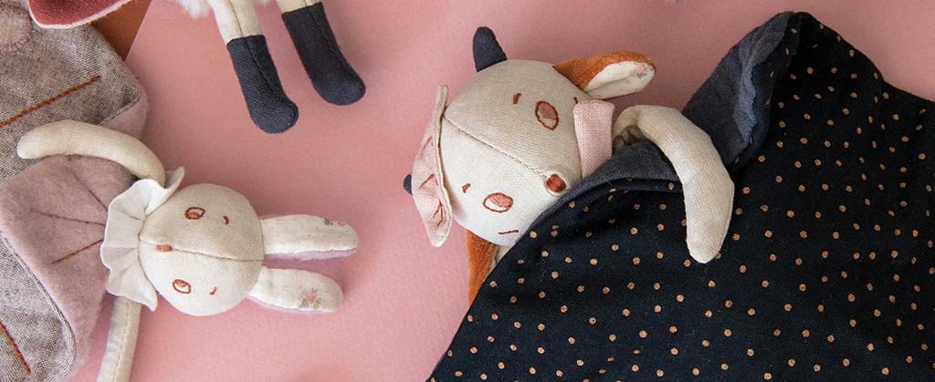 Soft handmade fabric animal plush toys lying on a pink background, featuring delicate stitching and vintage-style clothing.