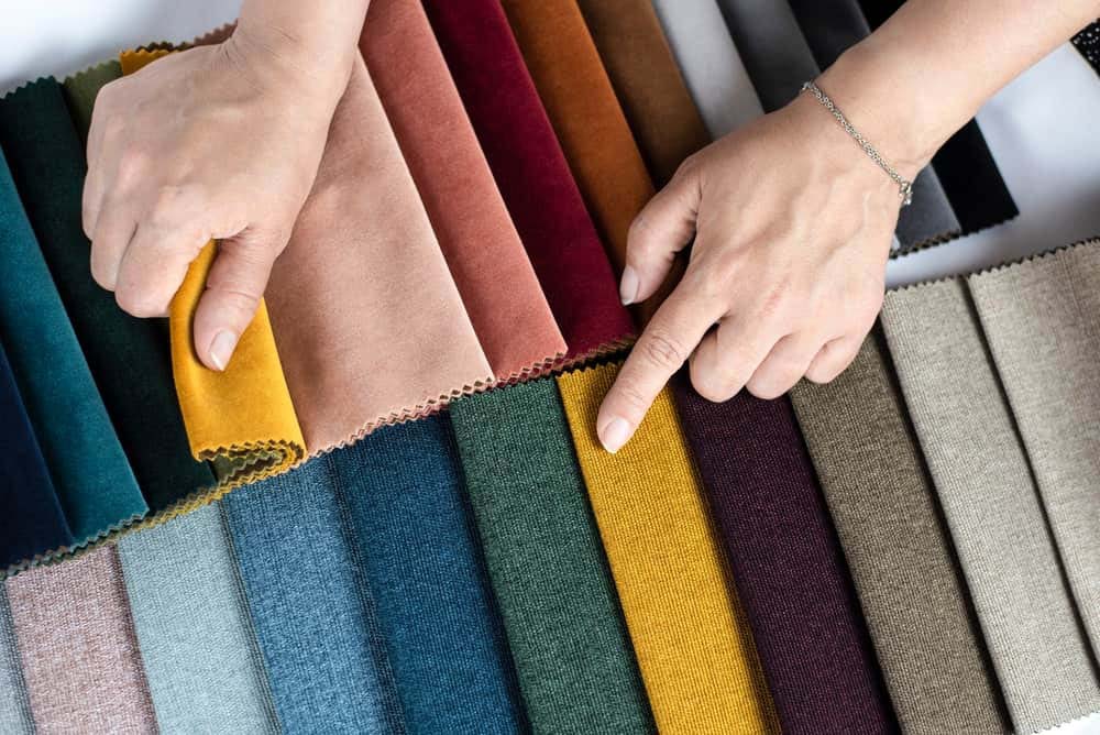 Close-up of two hands choosing from a variety of colorful fabric swatches arranged in rows, showing different textures and materials used for upholstery, crafts, or plush toy production.