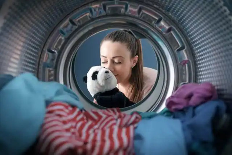 Inside view of washing machine with a woman reaching in to remove a clean panda plush toy surrounded by colorful laundry.
