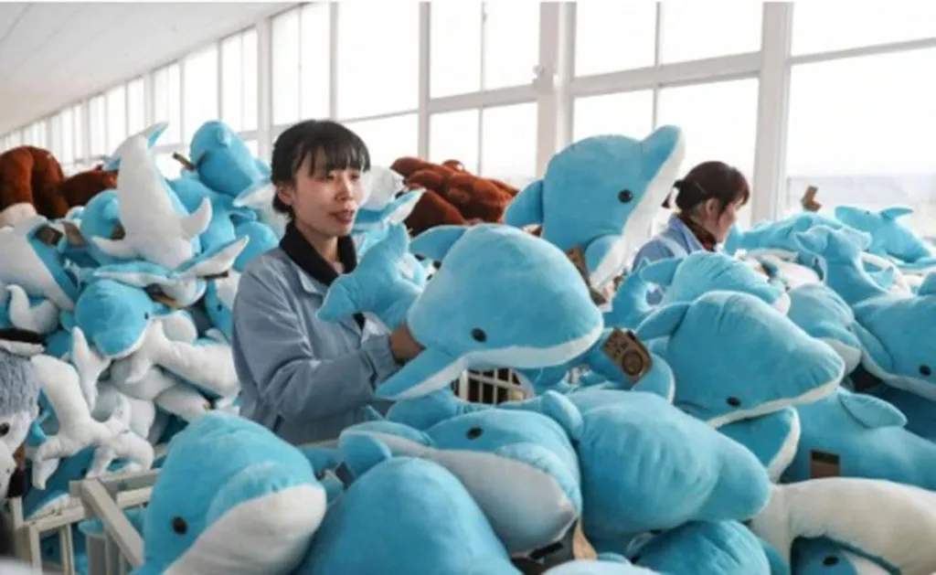 Workers inspecting and sorting blue dolphin plush toys inside a large plush toy manufacturing workshop with bins full of finished stuffed animals.