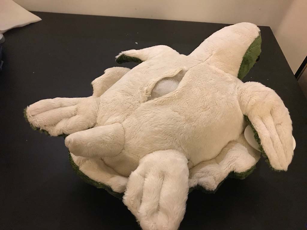 Damaged plush toy with a large torn seam on its back showing exposed stuffing, placed on a dark surface.