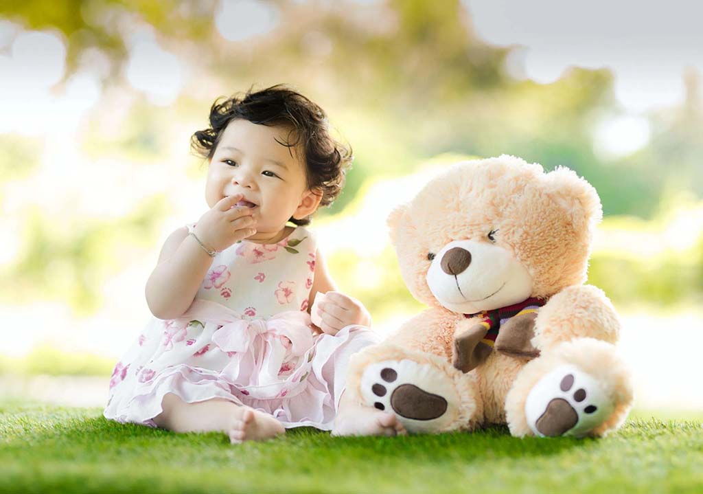 A smiling baby in a pink floral dress sits on the grass beside a large beige plush teddy bear, enjoying a bright and soft outdoor setting.