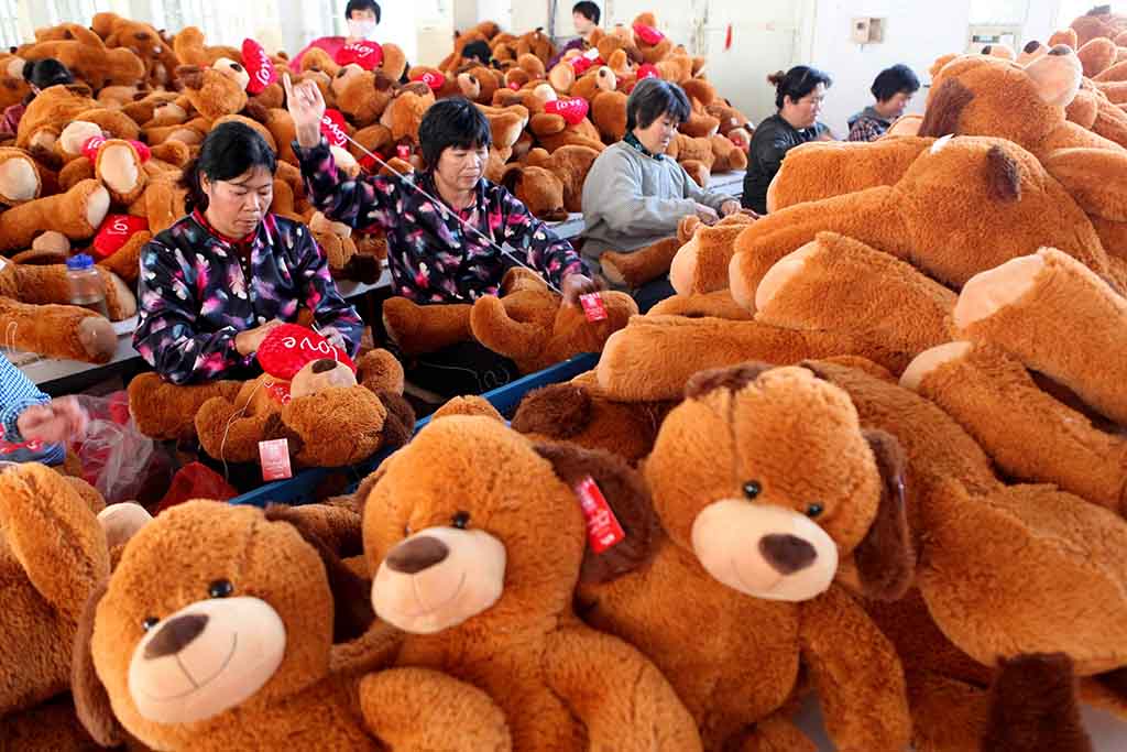 Workers in a plush toy factory sewing and assembling large brown teddy bears, surrounded by piles of stuffed bears in a production area.