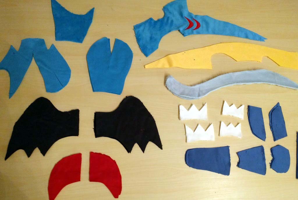 Assorted fabric pieces for a handmade plush toy laid out on a table, including wings, body shapes, fins, spikes, and accent parts ready for sewing and assembly.
