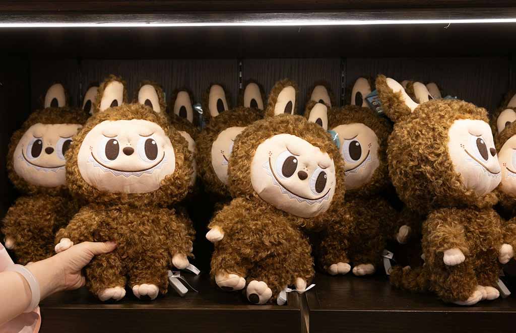 A shelf full of curly brown monster plush toys with big expressive faces and bunny-like ears, with a person’s hand picking up one of the plushies.