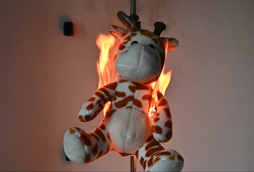 A stuffed giraffe toy undergoing a flammability test, showing controlled flames behind the plush to evaluate safety compliance for toy manufacturing standards.