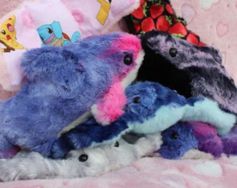 A pile of colorful plush sea-creature toys in shades of blue, purple, pink, and grey, featuring soft fur textures and small black eyes, arranged on a patterned pink background.