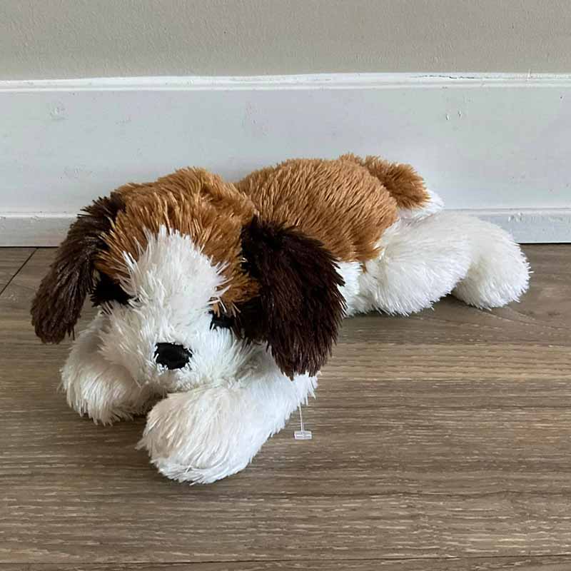 A soft, brown-and-white stuffed puppy lying flat on a wooden floor with floppy ears and a plush, shaggy texture.