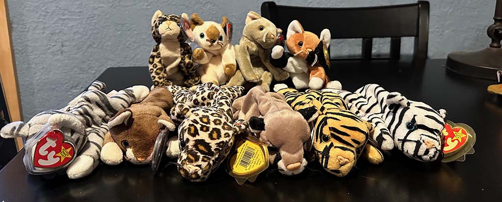 Row of assorted plush animal toys, including tigers, leopards, zebras, and other small stuffed animals, arranged on a black table with chairs in the background.